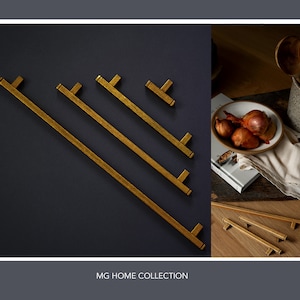 May include: A set of five gold-toned cabinet pulls in various sizes. The pulls are arranged in a diagonal line on a dark grey background. The text "MG HOME COLLECTION" is printed below the pulls.