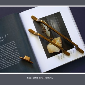 May include: Two gold-toned cabinet door pulls with a slightly curved design. The pulls are laying on top of an open magazine with the text "Power of Paint" visible. The magazine is on a dark grey surface.