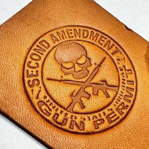 May include: A brown leather patch with a circular design featuring a skull and crossbones, two guns, and the text "Second Amendment Gun Permit".