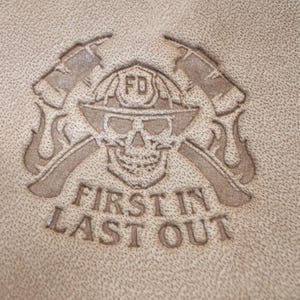 Puede incluir: Primer plano de un artículo de cuero marrón con un emblema de bombero en relieve. El emblema presenta una calavera con un casco con "FD", hachas cruzadas y el texto "FIRST IN LAST OUT".