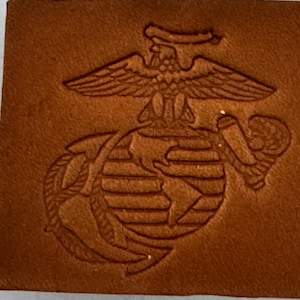 May include: A brown leather rectangle with an embossed emblem. The emblem depicts an eagle, globe, and anchor, a symbol of the United States Marine Corps. The leather has a textured finish and a warm, earthy tone.