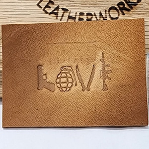 May include: A brown leather patch with a design of a gun, a grenade, and a knife. The design is embossed into the leather.