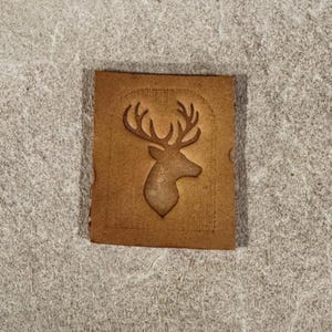 May include: A small, rectangular, tan leather patch featuring a debossed deer head with prominent antlers. The deer image is centrally placed on the patch, which has a slightly textured surface and rounded corners.