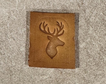 May include: A small, rectangular, tan leather patch with a debossed image of a deer head with large antlers. The deer is centered on the patch, which has a slightly textured surface and rounded corners.