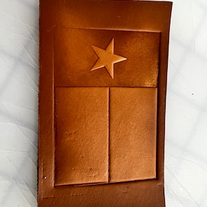 Puede incluir: Un parche de cuero marrón con una estrella blanca y dos líneas horizontales, que recuerda a la bandera del estado de Texas.