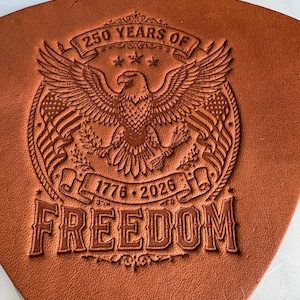 May include: A tan leather piece featuring a debossed design of a bald eagle with spread wings, two American flags, and the text 250 YEARS OF, 1776-2026, and FREEDOM. The eagle holds arrows and an olive branch within a circular border.