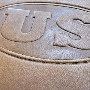 Puede incluir: Primer plano de un parche de cuero marrón con las letras "US" en relieve en un óvalo blanco.
