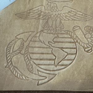 May include: A tan leather piece with a detailed carving of the United States Marine Corps emblem. The emblem features an eagle, globe, and anchor design, a symbol of the USMC.