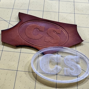 Puede incluir: Un sello de plástico transparente con las letras "CS" en forma ovalada. El sello se utiliza para crear una impresión en cuero.