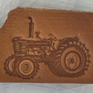Può includere: Un oggetto in pelle marrone con un'immagine in rilievo di un trattore d'epoca. Il trattore presenta grandi pneumatici posteriori, un camino e dettagli meccanici intricati. Il design generale è rustico e dettagliato.