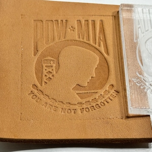 Op de afbeelding: Een bruine leren patch met een reliëfdesign van het POW MIA-logo. Het logo toont een silhouet van een soldatenhoofd, een wachttoren en de woorden "POW MIA" en "You Are Not Forgotten".