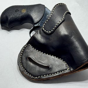 Könnte beinhalten: Eine schwarze Lederholster mit weißen Nähten, entworfen für einen Revolver. Die Holster hat eine gebogene Form und einen Ausschnitt für den Abzug. Der schwarze Griff des Revolvers ist sichtbar.