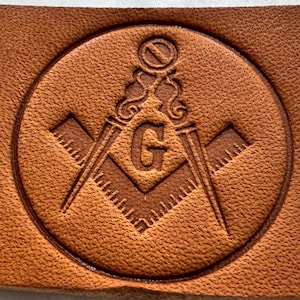 Könnte beinhalten: Nahaufnahme eines braunen Lederemblems mit einem Freimaurersymbol. Das Emblem zeigt einen Zirkel, ein Winkelmaß und den Buchstaben "G" in einem Kreis. Das Leder hat eine strukturierte Oberfläche.