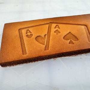 May include: A rectangular piece of tan leather with embossed playing card designs. The design features two Aces, one of hearts and one of spades. The leather has a rough, textured edge.