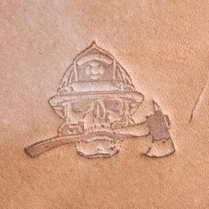 May include: A detailed, embossed design on a tan leather surface. The image features a skull wearing a firefighter's helmet, with an axe positioned across the skull. The design is a light brown color, creating a contrast against the lighter background.