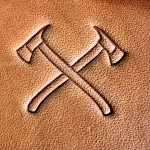 May include: Close-up of a tan leather surface with an embossed design of two crossed hammers. The hammers are detailed with precise lines and a textured appearance. The design is symmetrical, and the leather has a subtle, natural grain.