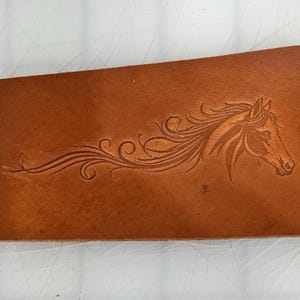 May include: Close-up of a brown leather piece featuring a detailed horse head design. The horse's mane flows gracefully, and the design is carved into the leather, creating a textured effect. The leather has a natural, warm tone.