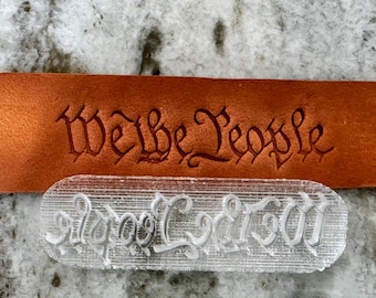 Könnte beinhalten: Ein transparenter Stempel aus Kunststoff mit den Worten "We the People" in einer dekorativen Schriftart. Der Stempel ist für die Lederbearbeitung gedacht.
