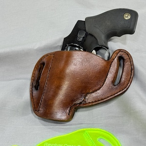 Könnte beinhalten: Ein schwarzer Revolver in einer braunen Lederholster. Die Holster hat eine gebogene Form mit zwei Schlaufen für einen Gürtel. Der Griff der Waffe ist schwarz und der Lauf ist silberfarben. Die Holster ist aus braunem Leder.