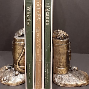 May include: A pair of gold-toned bookends shaped like golf bags with golf clubs sticking out of the top. The bookends are sitting on a textured base that resembles a rock. The bookends are holding up three books with the titles "of Whistler", "Centuries of Art Survey and Index", and "of Cezanne".