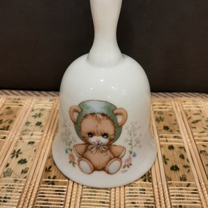 May include: White porcelain bell with a green and brown teddy bear design. The bear is wearing a green hat and has a brown bow. The bell has a white handle.