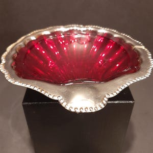 May include: A decorative, shell-shaped dish with a silver-coloured rim and a vibrant red interior. The rim features a beaded design. The dish is displayed on a black stand, showcasing its elegant form and colour contrast.