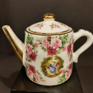 May include: White porcelain teapot with a gold-trimmed spout and lid. The teapot is decorated with pink and red roses and a pastoral scene. The lid has a gold knob. The handle is white.
