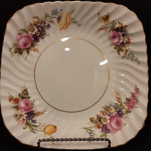 May include: A white square plate with a gold rim and a floral design. The plate has a raised center and a scalloped edge.