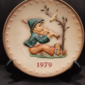 May include: A decorative plate featuring a young boy playing a flute, with a bird perched on a tree branch. The boy wears a green hat and jacket, and orange pants. The year "1979" is displayed in red. The plate has a cream background with star accents.