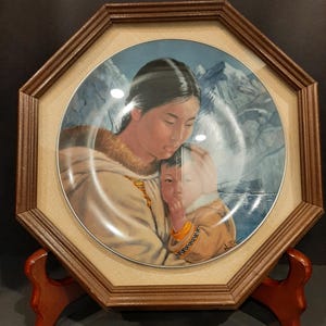 May include: Framed decorative plate featuring a mother and child with a mountain backdrop. The plate is set in an octagonal brown wooden frame with a tan fabric matting. The plate is displayed on a wooden stand.