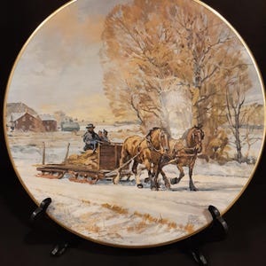 Op de afbeelding: Decoratieve plaat met een winterlandschap met een door paarden getrokken slee. De plaat heeft een gouden rand en een zwarte standaard. Het kunstwerk toont een landelijke omgeving met een grote boom en gebouwen op de achtergrond.