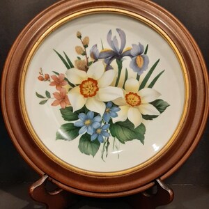 Könnte beinhalten: Ein dekorativer Teller mit einem Blumenstrauß aus weißen Narzissen mit orangefarbenen Zentren, blauen Schwertlilien und kleinen blauen Blumen. Der Teller ist in einem runden, dunkelbraunen Holzrahmen mit einem goldenen Innenrand gefasst. Der Teller wird auf einem Holzständer präsentiert.