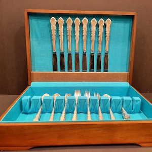 Könnte beinhalten: Eine Holzbesteckkiste mit türkisfarbenem Innenraum beherbergt ein Set aus silbernen Utensilien. Das Set umfasst Messer, Gabeln und Löffel, alle mit verzierten Griffen. Die Kiste ist geöffnet und zeigt das ordentlich angeordnete Besteck.