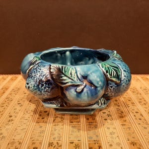 Pode incluir: Um vaso de cerâmica azul e verde em forma de tigela com desenhos de frutas em relevo. O vaso tem um acabamento esmaltado e é decorado com folhas e frutas.