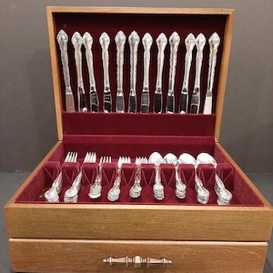 Peut inclure: Un coffret en bois pour couverts avec un intérieur en velours rouge, présentant un ensemble complet d'ustensiles en argent ornés. L'ensemble comprend des couteaux, des fourchettes et des cuillères, tous rangés dans le coffret.