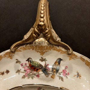 May include: A white porcelain serving dish with a gold rim and a gold handle. The dish is decorated with a floral pattern and birds.