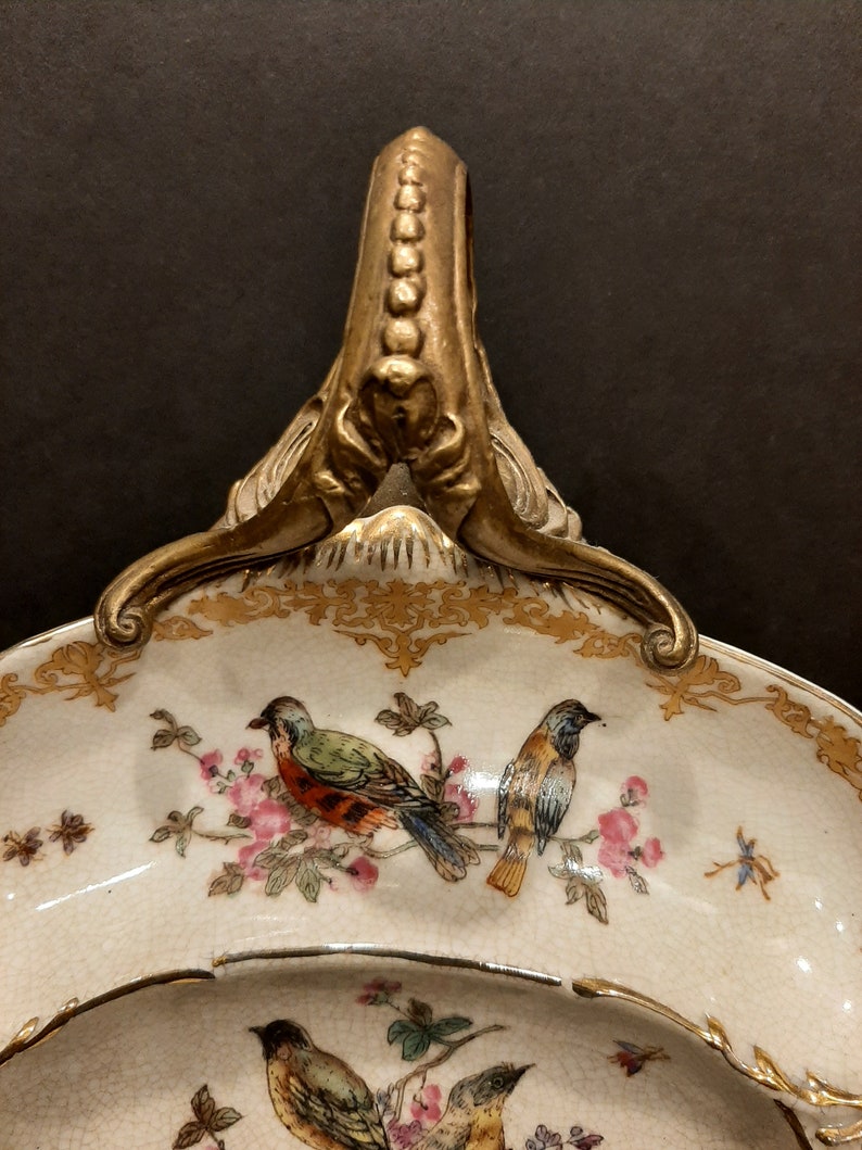 May include: A white porcelain serving dish with a gold rim and a gold handle. The dish is decorated with a floral pattern and birds.
