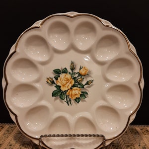 May include: A white ceramic egg plate with a scalloped rim and gold trim. The centre features a floral design of yellow roses and green leaves. The plate has 12 egg-shaped indentations.