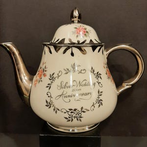 Könnte beinhalten: Cremefarbene Teekanne mit silbernen Akzenten, mit floralem Design und dem Text "Silver Wedding Anniversary 25 years". Die Teekanne hat einen gebogenen Ausguss, einen Henkel und einen Deckel mit silberner Spitze. Das florale Design umfasst rosa und orange Rosen.