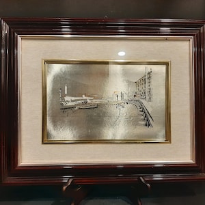 May include: Framed artwork featuring a silver-toned landscape scene with buildings, a bridge, and boats. The artwork is set within a gold-toned frame, surrounded by a beige mat, and encased in a dark brown wooden frame. The piece is displayed on a wooden stand.