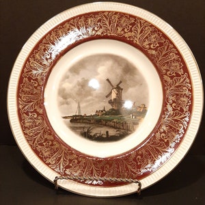 May include: A white ceramic plate with a red and gold border. The plate features a vintage illustration of a windmill in a rural landscape.