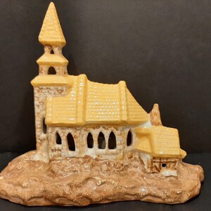 May include: A ceramic church figurine with a yellow roof and brown stone details. The church has a tall steeple and a small side building. The figurine is sitting on a brown base.