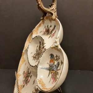 May include: A white ceramic serving dish with three sections, featuring a gold trim and painted bird designs. The dish has a decorative brass handle.
