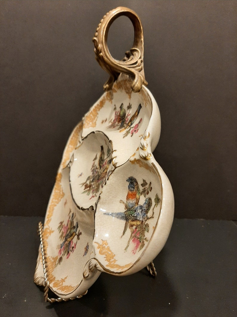May include: A white ceramic serving dish with three sections, featuring a gold trim and painted bird designs. The dish has a decorative brass handle.