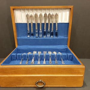 Peut inclure: Coffre à couverts en bois avec intérieur en velours bleu. Le coffre contient un ensemble de couteaux, fourchettes et cuillères argentés. Le coffre a une poignée en laiton et est ouvert, révélant les couverts.