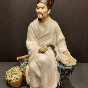 May include: A glazed ceramic figurine of a seated scholar wearing a white robe and a dark brown cap. The figure has a goatee, holds a book in one hand, and sits on a textured blue and brown base next to a woven basket filled with green foliage.