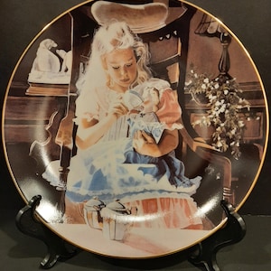 May include: Decorative plate featuring a young girl with long blonde hair, dressed in a light pink dress, holding a doll. The plate has a gold rim and depicts a scene with a chair, flowers, and a tea set. The plate is displayed on a black stand.