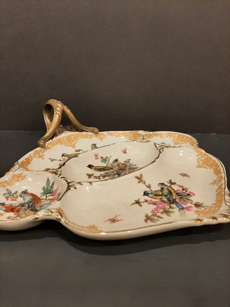 May include: A white ceramic serving tray with a gold rim and a gold handle. The tray is divided into four sections and features a floral design with birds.