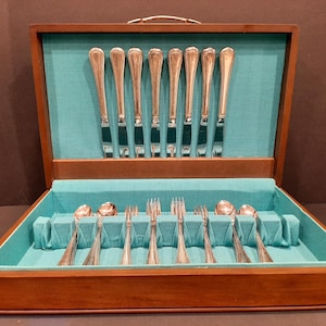 Könnte beinhalten: Eine Holzbesteckkiste mit türkisfarbenem Innenraum, die ein Set aus silbernen Utensilien enthält. Das Set umfasst Messer, Gabeln und Löffel, die ordentlich in den Fächern angeordnet sind. Die Kiste ist geöffnet und zeigt den Inhalt, und hat einen Griff oben.