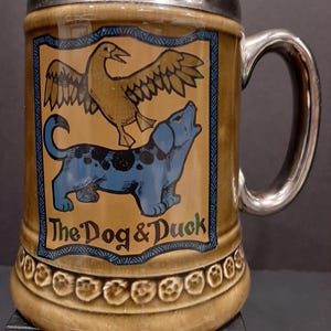 May include: A brown ceramic mug with a silver lid and handle. The mug features a colorful illustration of a dog and a duck with the text "The Dog & Duck".